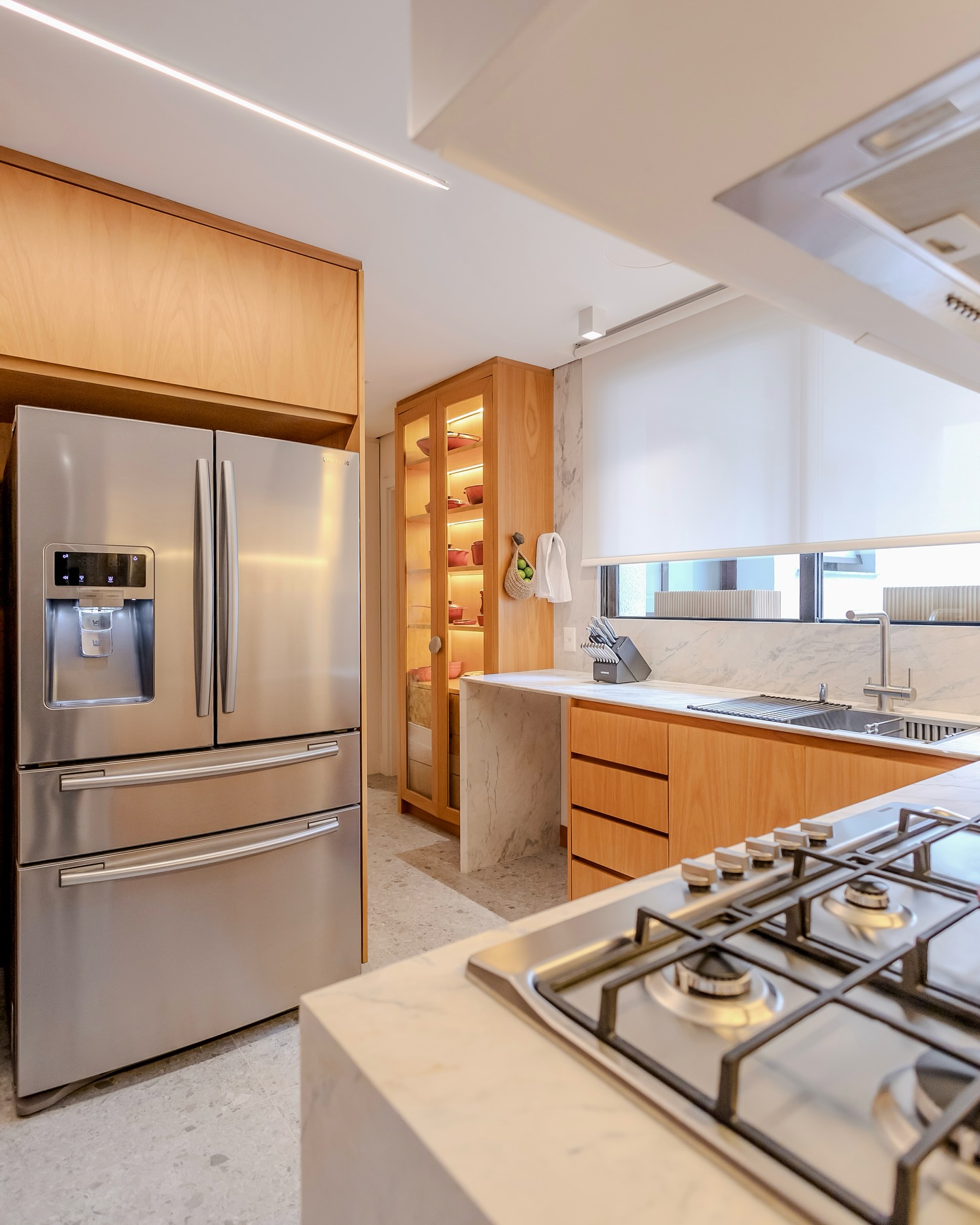 Cozinha planejada moderna com armários em madeira natural, bancada de mármore, despensa integrada com iluminação LED e eletrodomésticos de inox embutidos